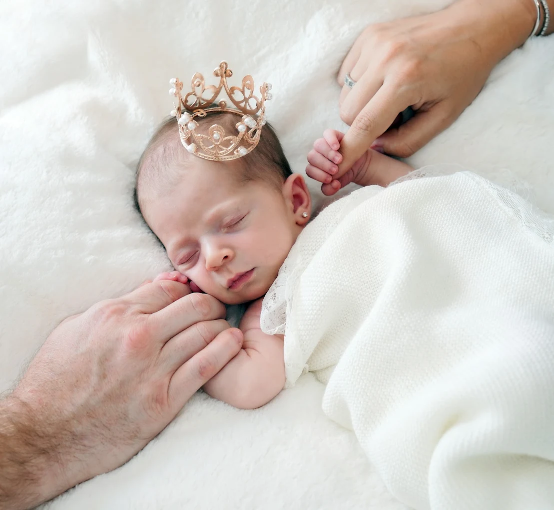 Newborn baby wearing a tiny crown while holding mom and dad's hands during a photography session at Adrisabel Photography